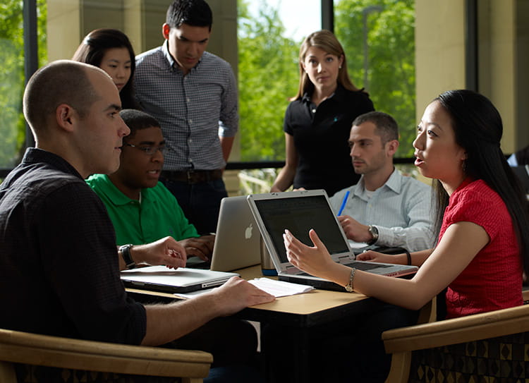A group of seven students gather around two laptops to work together.