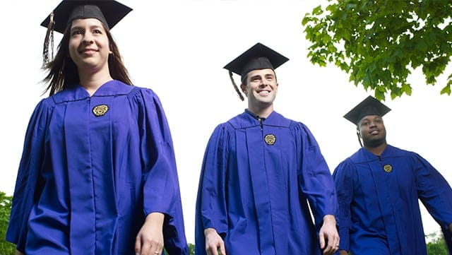 Three graduates dressed in regalia stand tall against a clear sky.