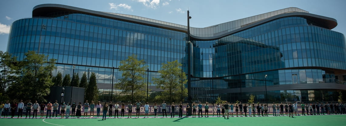 Men and women wearing Kellogg shirts lined up side-by-side in front of the Kellogg Global Hub. The glass building reflects the blue sky and few clouds.