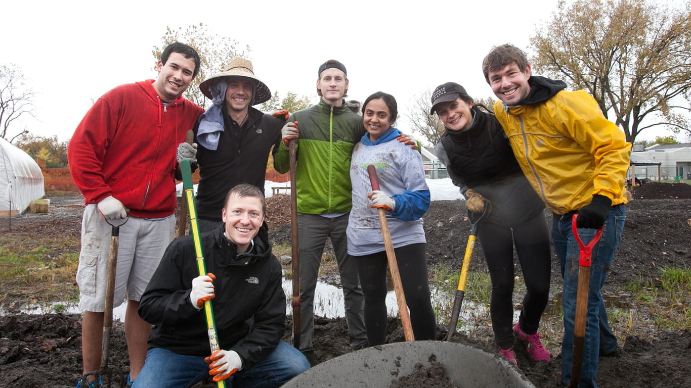 Seven smiling individuals holding shovels while wearing fall attire outdoors