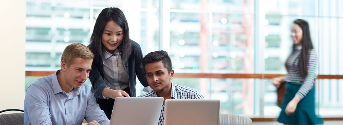 A group of three students have a conversation while pointing at laptops.