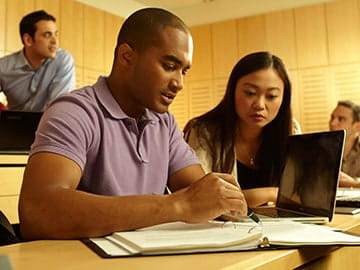 Two young students work together on a laptop in a lecture hall.