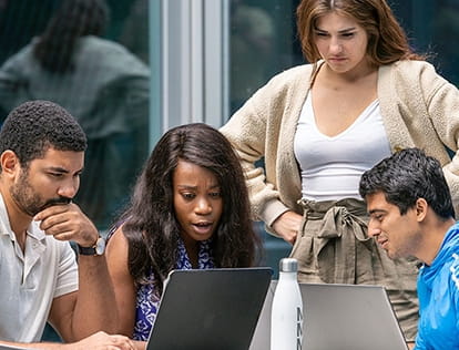 Four Kellogg students of varying ethnicities and genders collaborate at two laptops, on a sunny day beside a Kellogg Global Hub glass wall