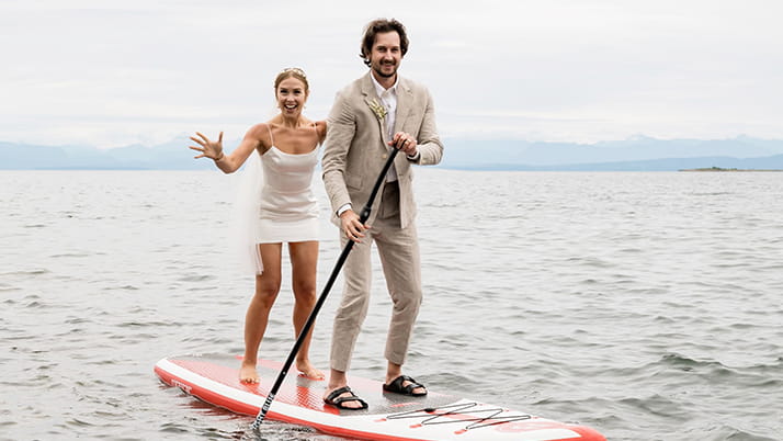Kellogg student Alex Tyrrell on her wedding day, with her husband. Both are wearing formal attire and riding a paddleboard on a lake.