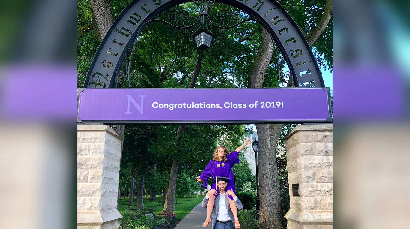Michelle Wright wearing purple robes on graduation day, sitting on a friend's shoulders underneath the Northwestern arch