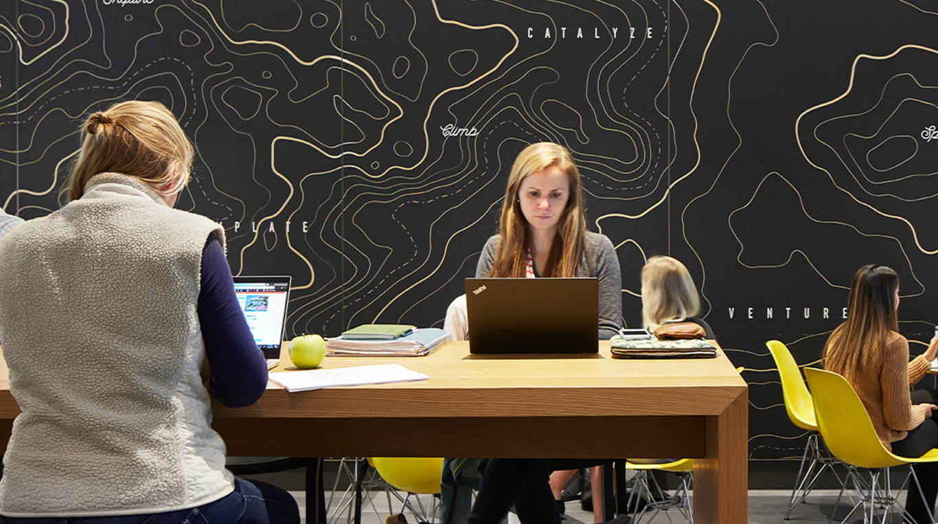 A woman sits at a cafe at the Global Hub, working on a computer against a black and yellow backdrop.