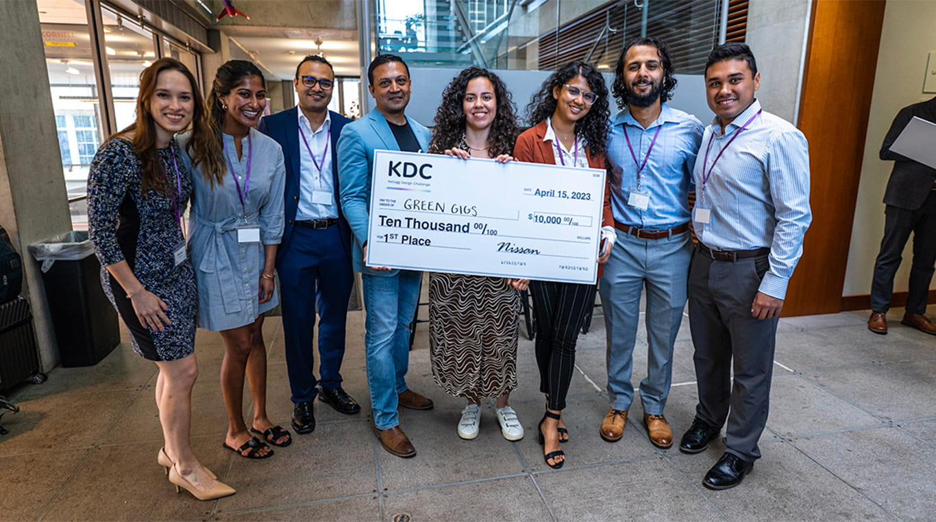 Eight people, including the Green Gigs student team, stand and smile posing with a large check indicating they won first place and $10,000 in the Kellogg Design Challenge.