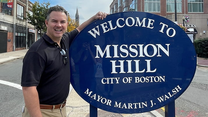 Mission Hill Capital founder Greg Kennealey ’03 MBA poses by a round blue sign with white lettering that reads "Welcome to Mission Hill, City of Boston"