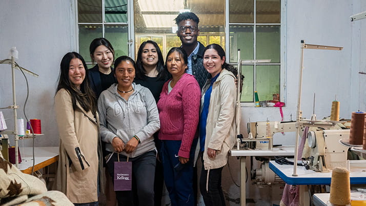 Students stand with a seamstress in her workspace, surrounded by sewing machines and items