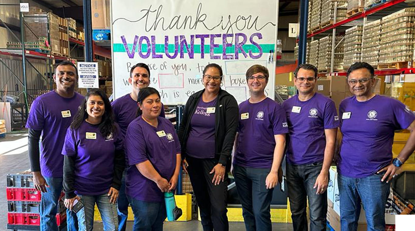 Kellogg alumni volunteers wearing purple shirts stand in a warehouse below a sign that says "Thank you volunteers"