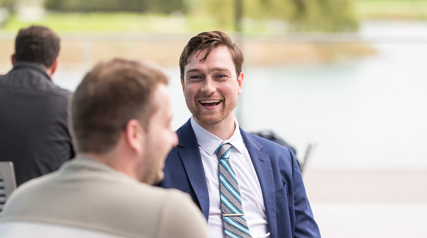 A Kellogg student in a navy suit and striped tie sits outside on a bench and talks with his classmate