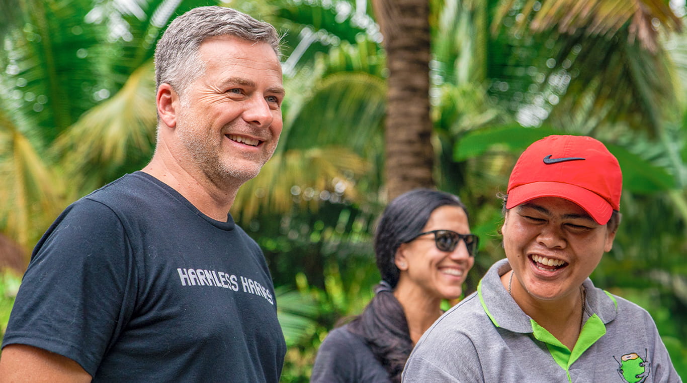 Kellogg graduate Ben Mand wears a black shirt with the Harmless Harvest logo while outdoors in a tropical area with a group of people