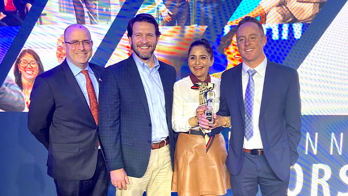 Indra Sandal stands on stage in a group of people against a bright video backdrop, holding a statuette at an awards ceremony.