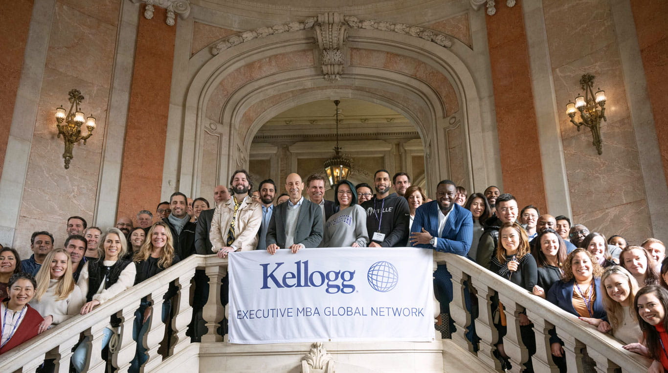 A large group of Kellogg Executive MBA students stand on a staircase at an historic Lisbon building, with a banner reading "Kellogg Executive MBA Global Network"