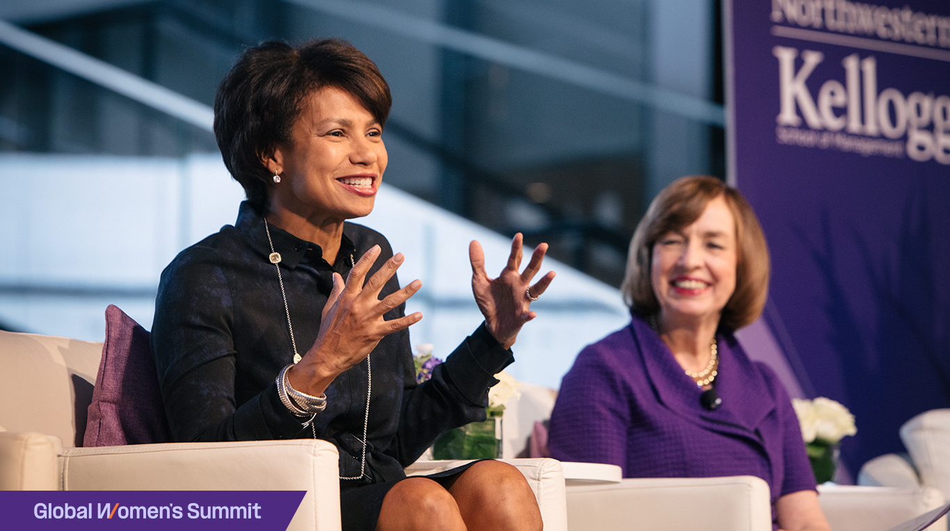 Two women sit side by side on a stage giving a panel discussion. A purple banner with the Kellogg logo is in the background.
