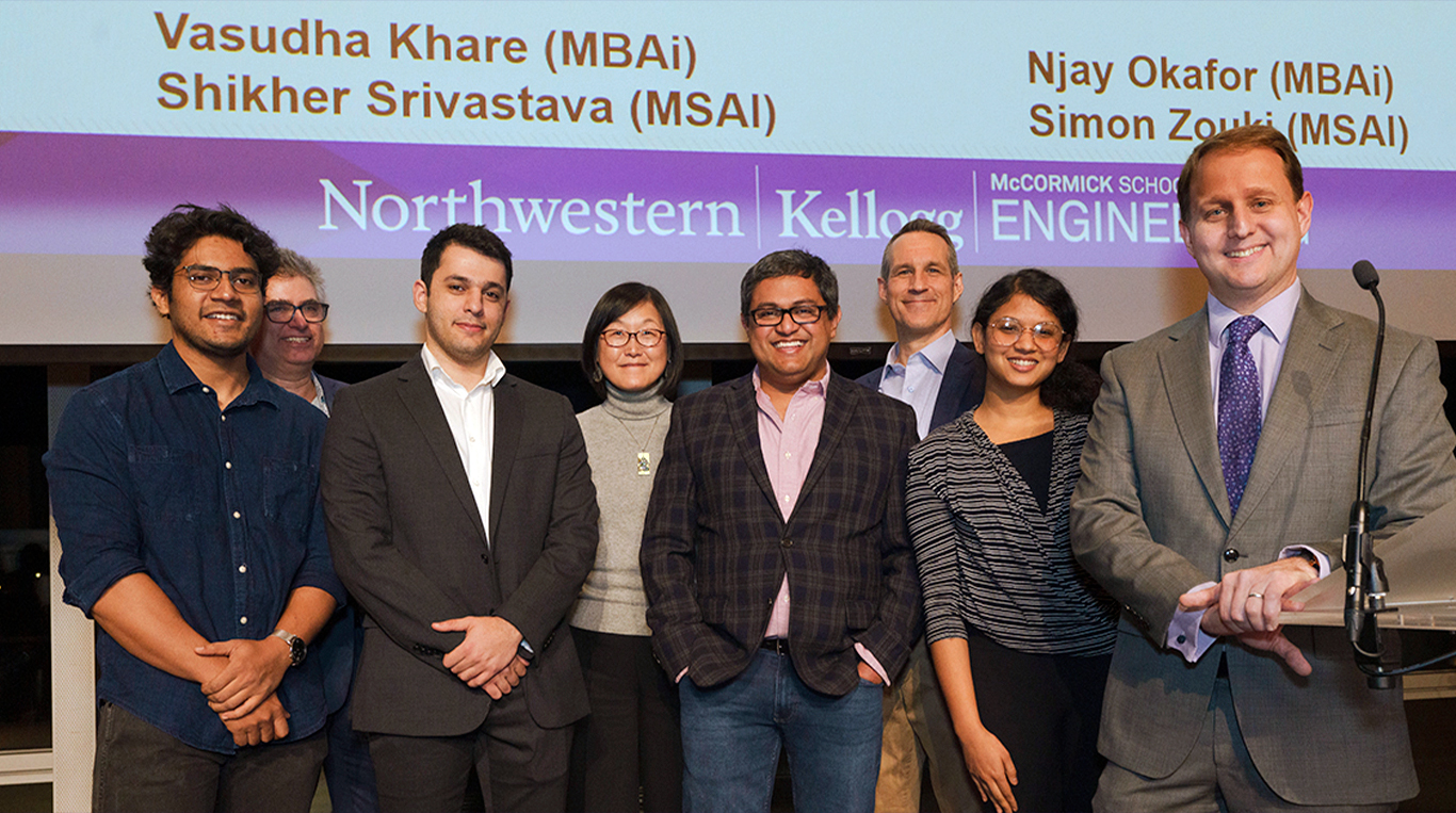 MBAi and MSAI students and faculty from Northwestern's Kellogg School of Management and McCormick School of Engineering pose together on stage after a student project showcase. A presentation slide above them displays several students’ names.