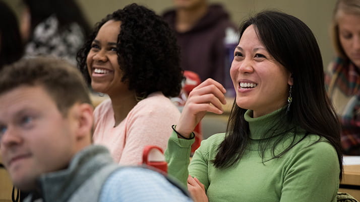 A Kellogg student in a green turtleneck smiles while sitting in a lecture hall surrounded by classmates