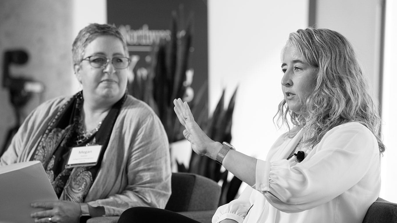 Faculty members Meghan Busse and Megan Kashner sit side by side on a stage giving a talk; Busse speaks while Kashner looks on.
