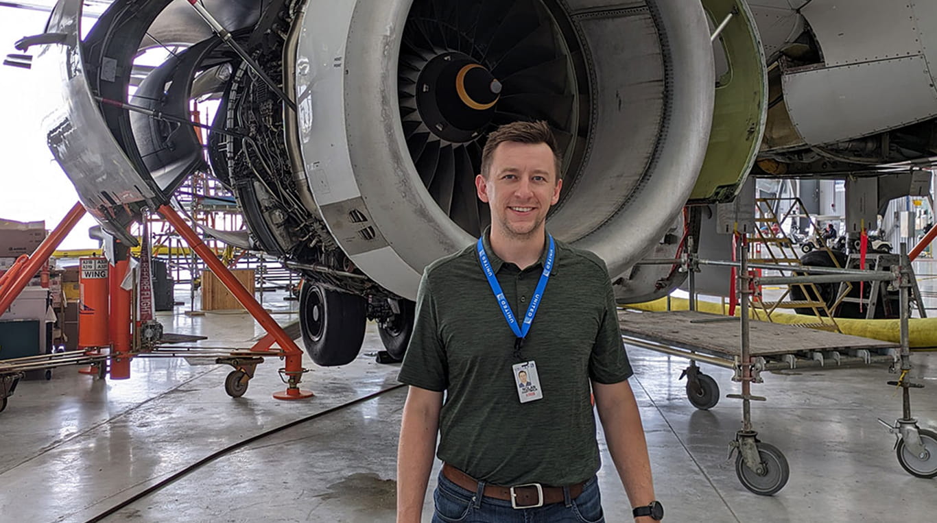 MBAi graduate Nolan Hartwick wears a blue United lanyard and stands in front of a commercial aircraft under construction in an airplane hangar. The plane's engine is visible behind him.