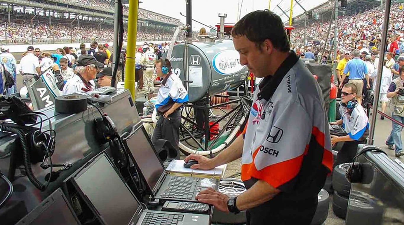 A pit crew member of a car racing team stands in front of a bank of laptops, at a racetrack. He wears a team jersey and behind him are a crowd of other pit crew members and spectators.