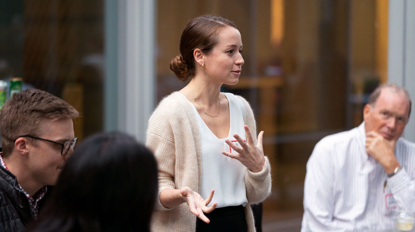 A Kellogg student wearing a white shirt, black pants and tan cardigan gestures as she speaks to classmates.