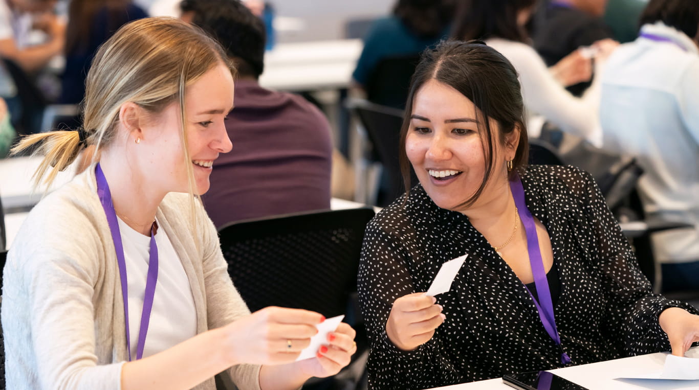 Two women, both Kellogg MBA students, laugh and smile as they participate in a conversation and show each other notecards. Both are wearing purple lanyards.