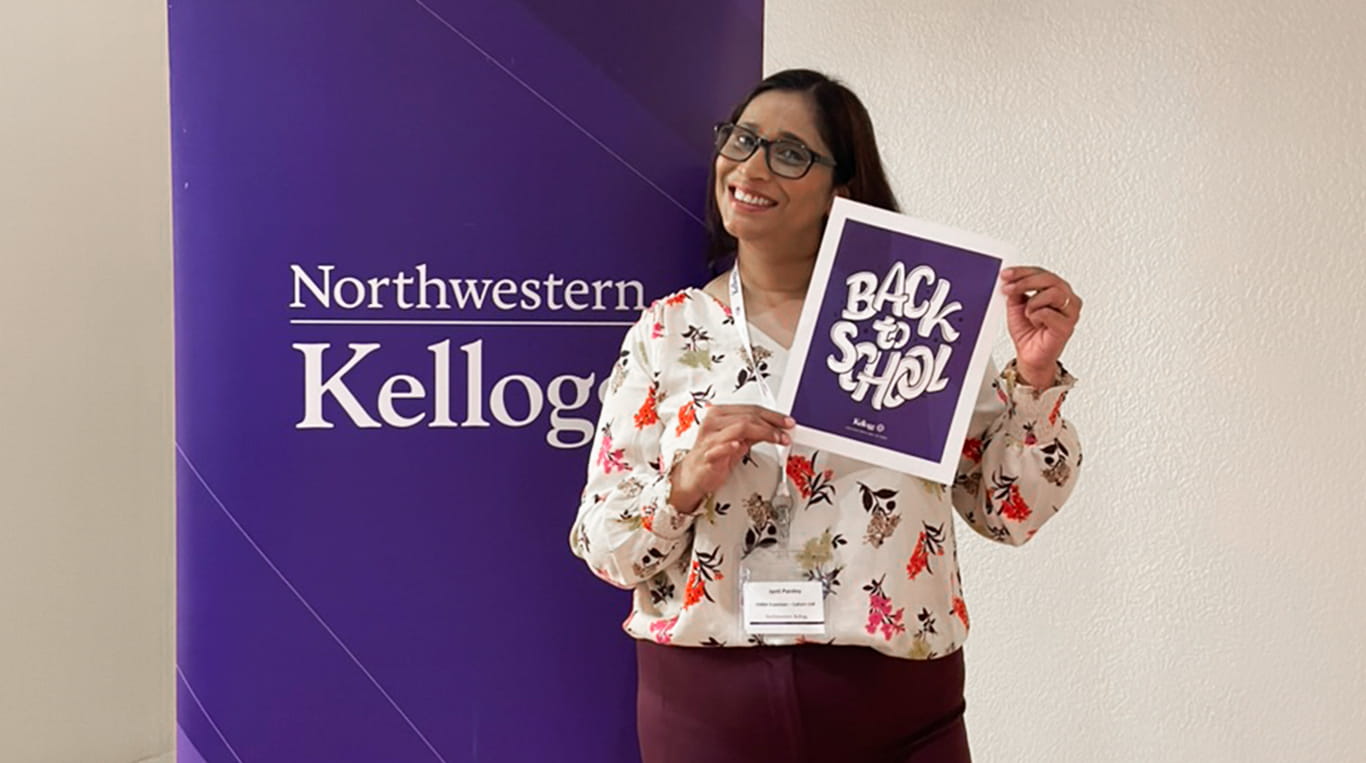 EMBA student Jyoti Pandey smiles while holding a sign that reads 'Back to School' in bold white lettering against a purple background. She stands in front of a large purple banner with the Northwestern Kellogg logo. The woman is wearing glasses, a floral blouse, and a name tag, standing in a bright indoor setting.