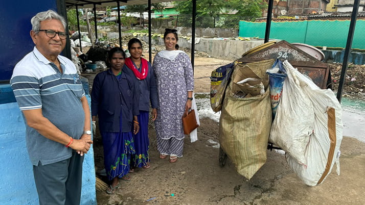 A group of four people including EMBA student Jyoti Pandey stand outside near a waste collection area, next to a garbage cart loaded with large sacks.