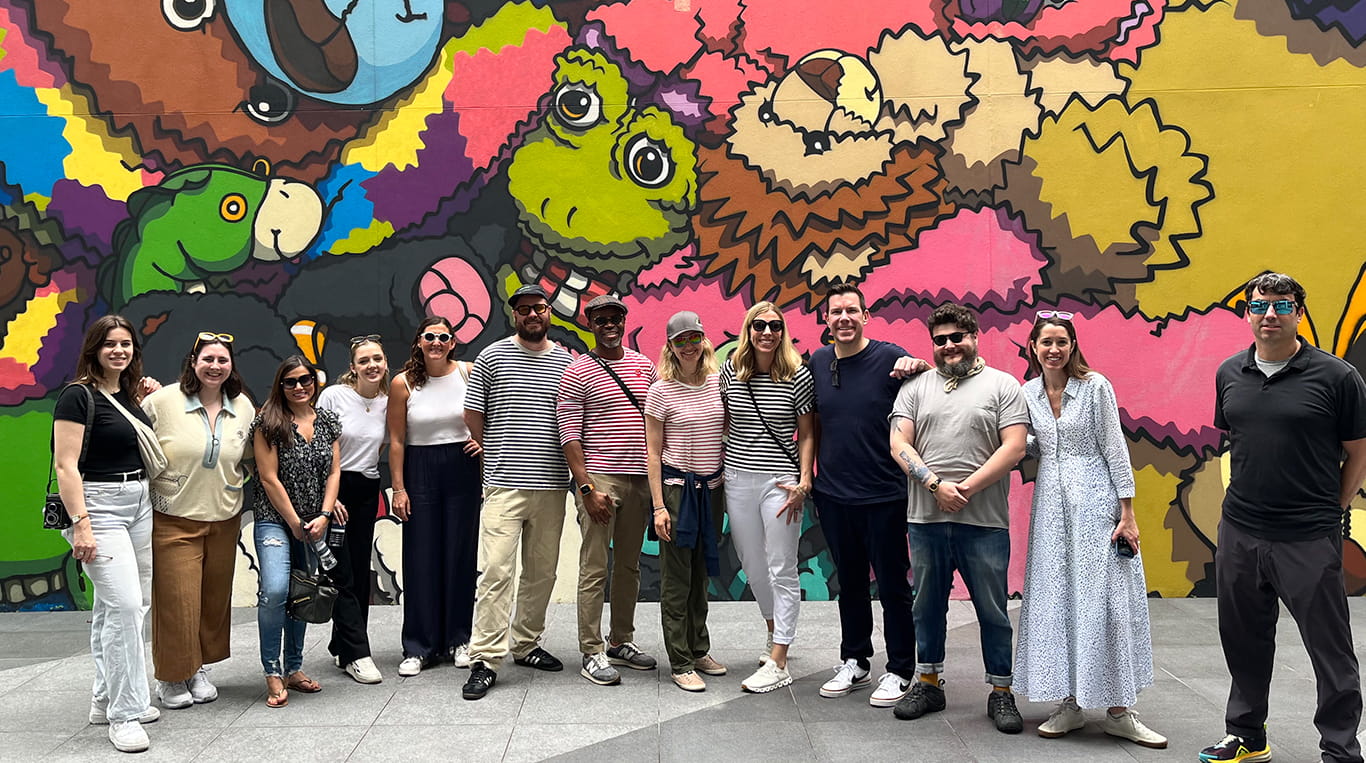 The Episode Four team stands smiling in front of a vibrant, colorful mural featuring cartoon-like animal characters, during a recent group outing. The group, casually dressed, includes men and women with a mix of sunglasses and summer attire. 