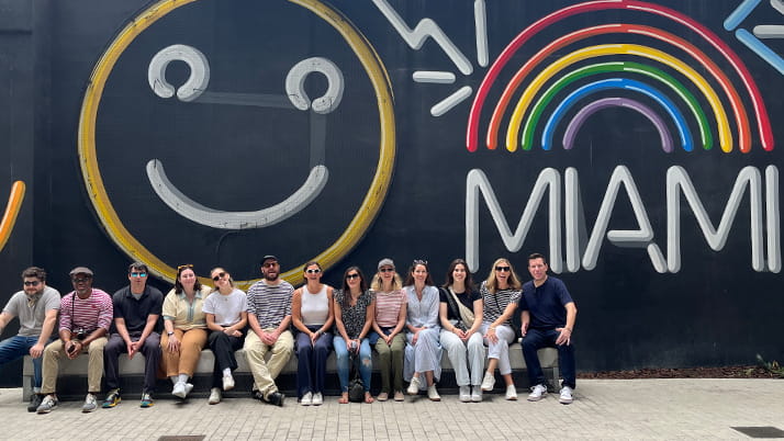 The Episode Four team sits together in front of a wall mural featuring a large, smiling face on the left and a colorful neon rainbow over the word MIAMI on the right. The group members are casually dressed and seated side by side, smiling and enjoying the bright, cheerful background.