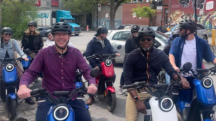 A group of individuals on electric scooters, wearing helmets, ready to explore an urban area. They appear excited and prepared for an adventure, with smiles and a mix of colorful scooters. 