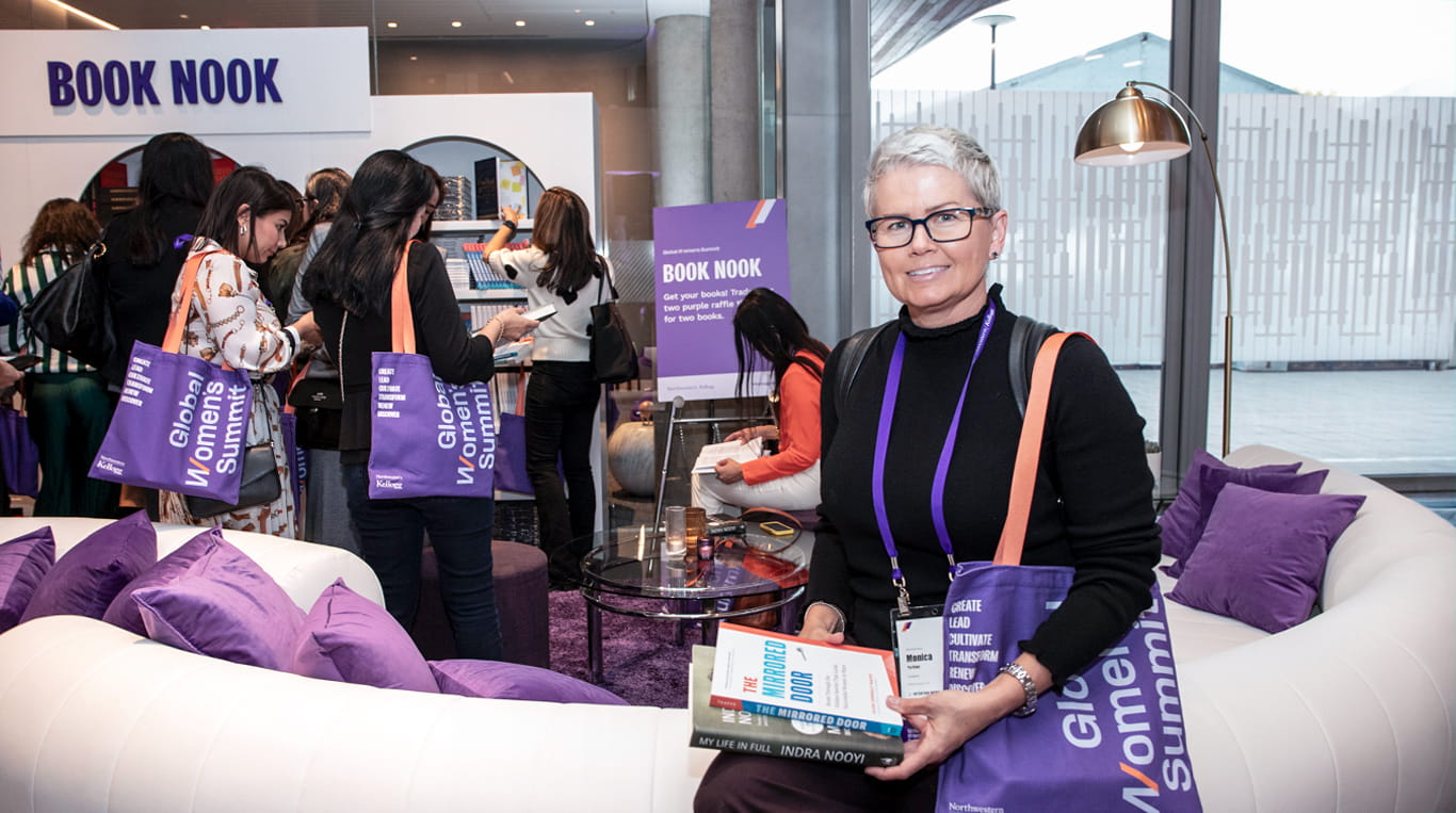A woman holds a stack of books at the 'Book Nook' area during the Kellogg Global Women's Summit. She is surrounded by other attendees, some browsing bookshelves and others with branded purple tote bags. The scene exudes a vibrant and welcoming environment, with purple decor accents and cozy seating arrangements.