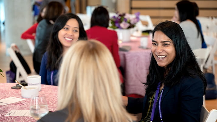 A group of professional women engage in lively conversation around a round table at the Kellogg Global Women's Summit. Smiling and appearing relaxed, they enjoy each other's company in a welcoming networking environment. The table is decorated with a pink tablecloth, floral centerpiece, and coffee mugs, creating an inviting atmosphere.