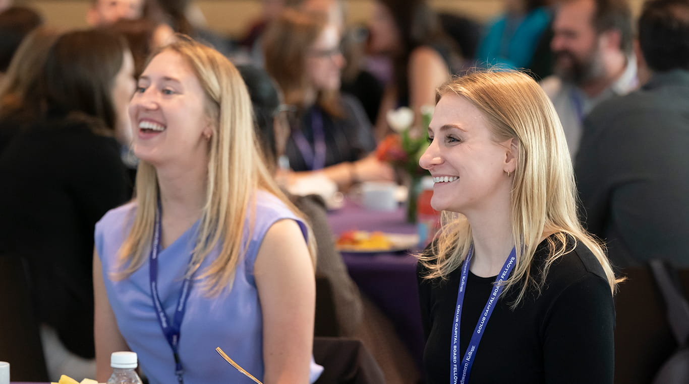 Two women smile and laugh while seated at a table during a Kellogg School of Management event. They wear lanyards and appear engaged and delighted, suggesting an atmosphere of camaraderie and positive energy. The background shows other attendees in similar cheerful interactions, with tables decorated with floral centerpieces.
