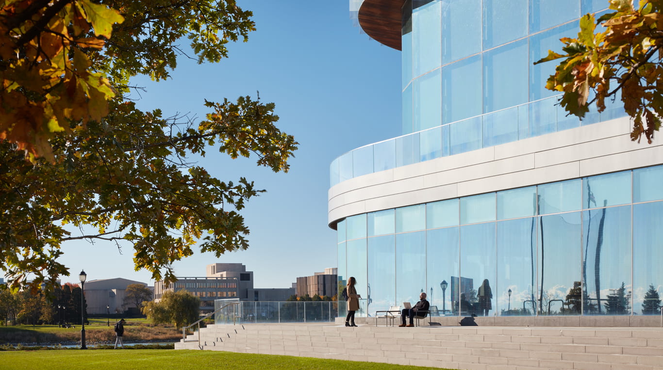A scenic view of the Kellogg School of Management campus. The glass-walled Global Hub building shines under a clear blue sky, with individuals engaging on the steps outside. A tree with autumn foliage frames the scene, adding vibrant orange and yellow tones. In the distance, Northwestern University's other buildings are visible across the green expanse of lawn.