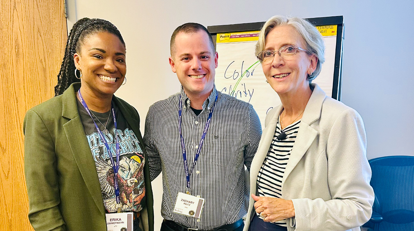 Three individuals smile at the camera in a professional setting. They are wearing conference badges and standing in front of a flip chart with colorful handwritten notes. 