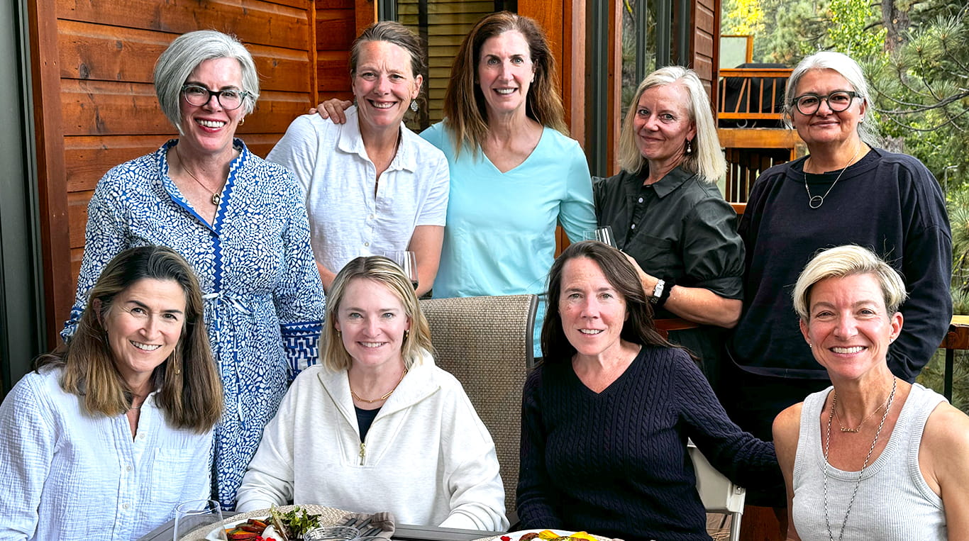 A group of nine women gathers around an outdoor dining table on a cozy wooden patio. Four women are seated with plates of food, while five stand behind them, smiling warmly. The setting suggests a casual, intimate gathering, with a backdrop of rustic architecture and greenery.