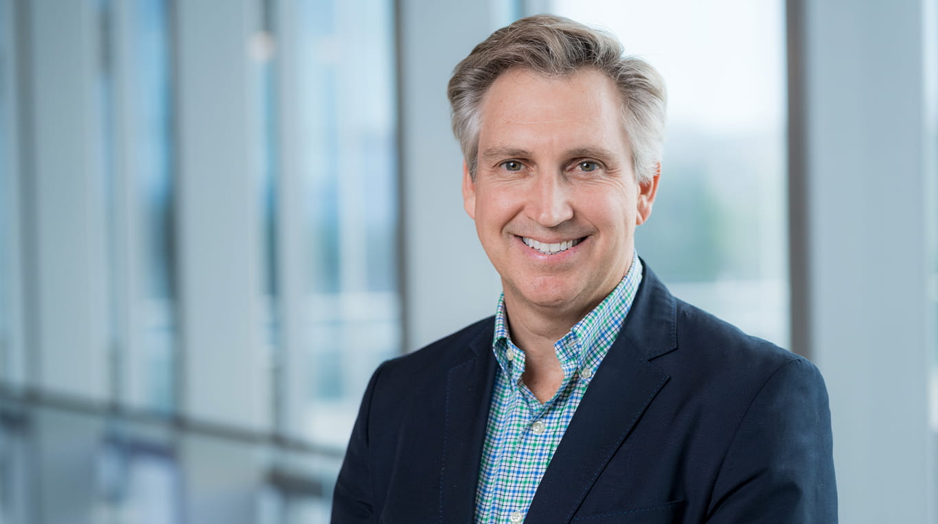 Kellogg alumnus Kent Hawryluk smiles while standing in the Kellogg Global Hub in Evanston. He wears a navy blazer and a light blue checkered shirt.