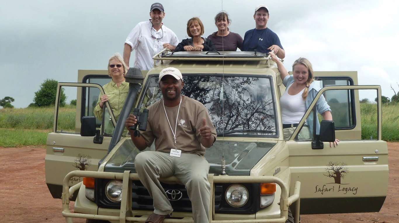 A group of six people pose with a safari guide in front of a beige Safari Legacy vehicle on a dirt road, surrounded by lush greenery. The safari guide, seated on the vehicle’s front bumper, smiles while holding binoculars, while the others stand on the vehicle’s running boards and roof rack, dressed in casual outdoor attire.