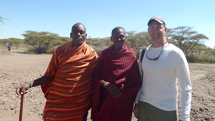 Three people standing together in an arid landscape with scattered acacia trees in the background. Two individuals are wearing traditional Maasai wraps - one in bright orange/red striped fabric and the other in solid red patterned fabric. The person on the left holds a walking stick. The third person on the right is wearing a white long-sleeved shirt, olive pants, and a cap.