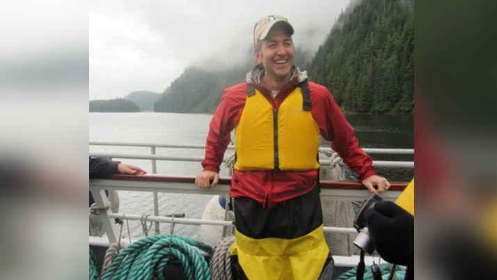 A person standing on the deck of a boat, wearing a bright yellow life vest over a red jacket and yellow-and-black waterproof pants. They are smiling at the camera while standing beside the boat railing. In the background is a misty fjord or inlet with steep, forested mountains partially obscured by low clouds.