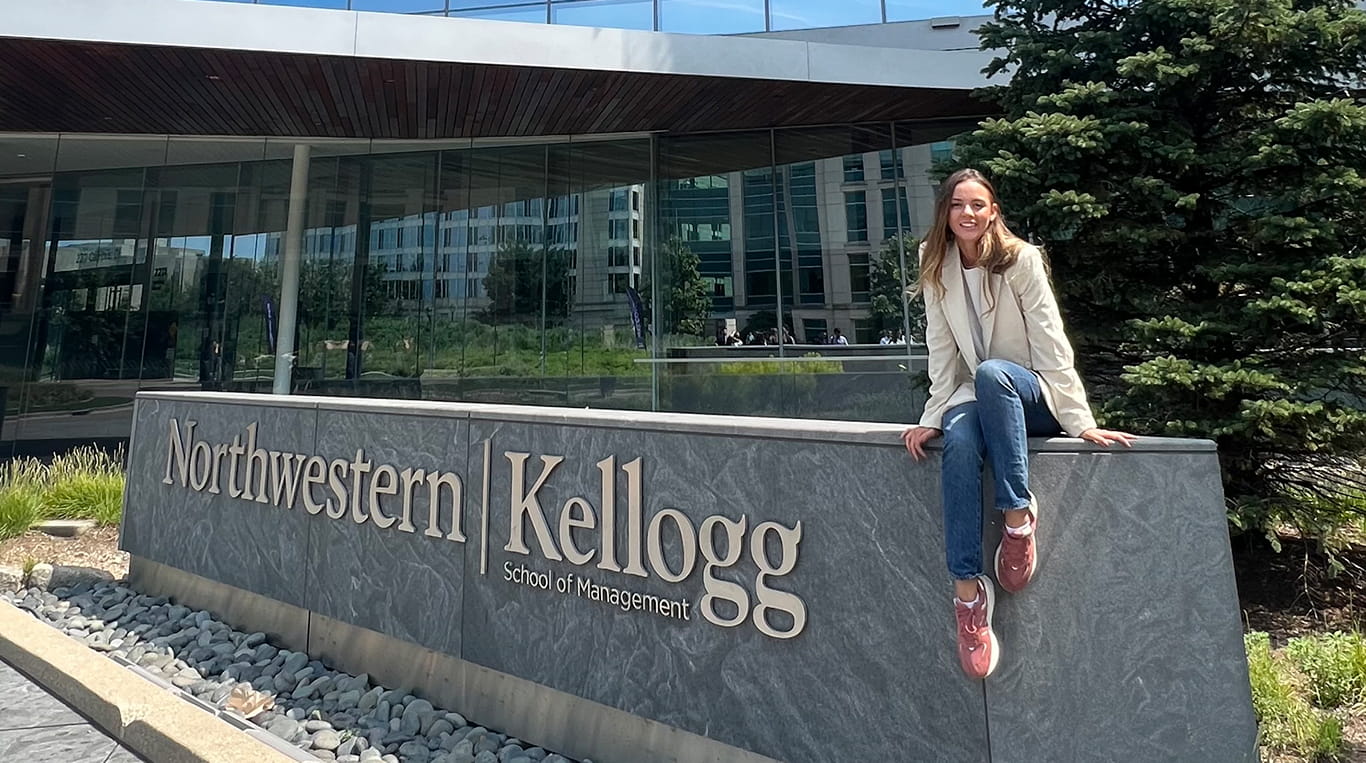 A young woman with long brown hair sits on a stone sign that reads "Northwestern | Kellogg School of Management." She is wearing a cream-colored blazer, blue jeans, and pink sneakers, smiling at the camera. The Kellogg Global Hub building and greenery are visible in the background.