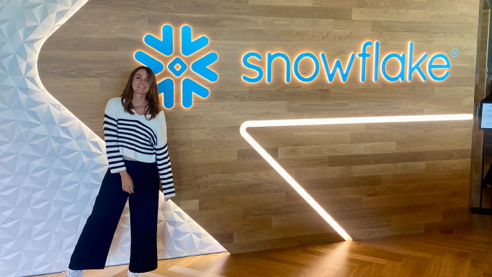 A young woman with shoulder-length brown hair stands smiling in front of a wooden wall featuring the illuminated Snowflake company logo. She is wearing a white and navy striped sweater, dark blue wide-leg pants, and white sneakers. The modern office setting includes geometric design elements and LED accent lighting.