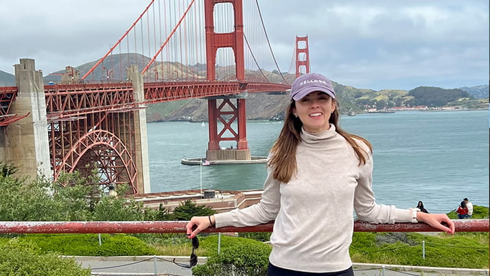 A young woman wearing a light beige turtleneck sweater and a purple cap with "Kellogg" embroidered on it stands in front of the Golden Gate Bridge in San Francisco. She is smiling, holding sunglasses in one hand, and resting her other arm on a red railing. The bridge, water, and hills are visible in the background under a cloudy sky.