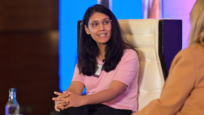 Roshni Nadar Malhotra ’03, ’08 MBA, Chairperson, HCLTech speaks with Dean Francesca Cornelli a the Pragati conference. She wears a pink shirt and glasses and sits on a white leather chair, speaking and gesturing with her hands during a panel discussion. The background features a blue-lit stage, creating a professional and dynamic setting. A bottle of water sits on a nearby table.