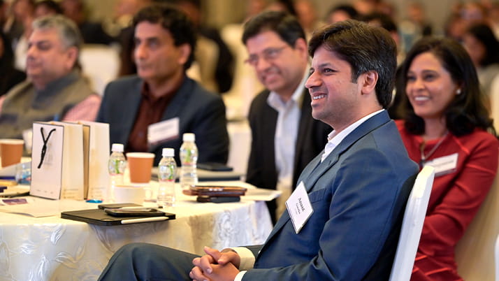 Attendees at the Pragati conference, seated at tables covered with white tablecloths. In the foreground, a smiling man in a blue suit with a name badge is prominently featured. To his right is a woman in a red outfit who is also smiling. Behind them are several other attendees in business attire, some wearing name badges. On the tables are water bottles, coffee cups, and what appear to be conference materials or gift bags.