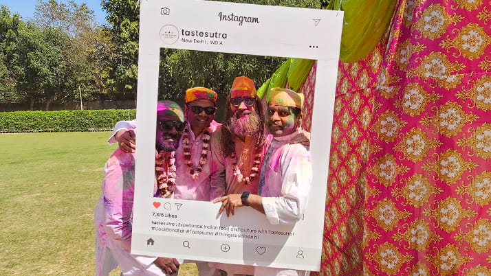 Four TechVenture India students celebrating Holi (the Indian festival of colors) posing behind a large Instagram frame prop. The Executive MBA students are covered in pink and orange colored powder and wearing flower garlands around their necks. 