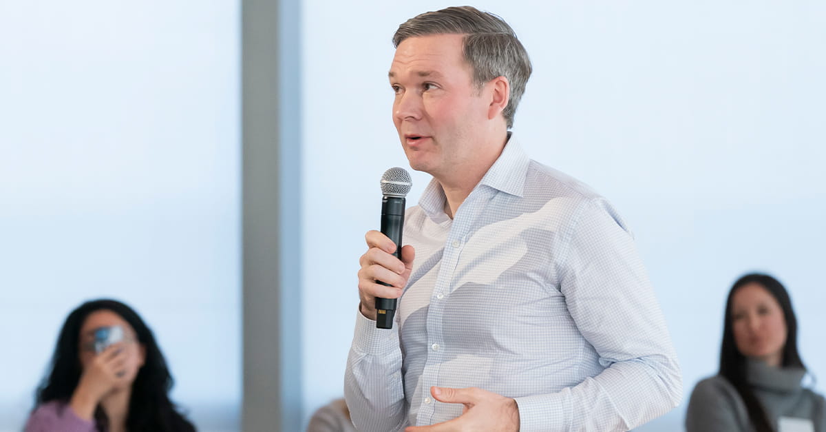 Kellogg alum Matt Peterson holding a microphone and speaking during an event at the Kellogg Global Hub. He is wearing a light blue checkered shirt and is framed in soft natural light, with blurred attendees visible in the background.