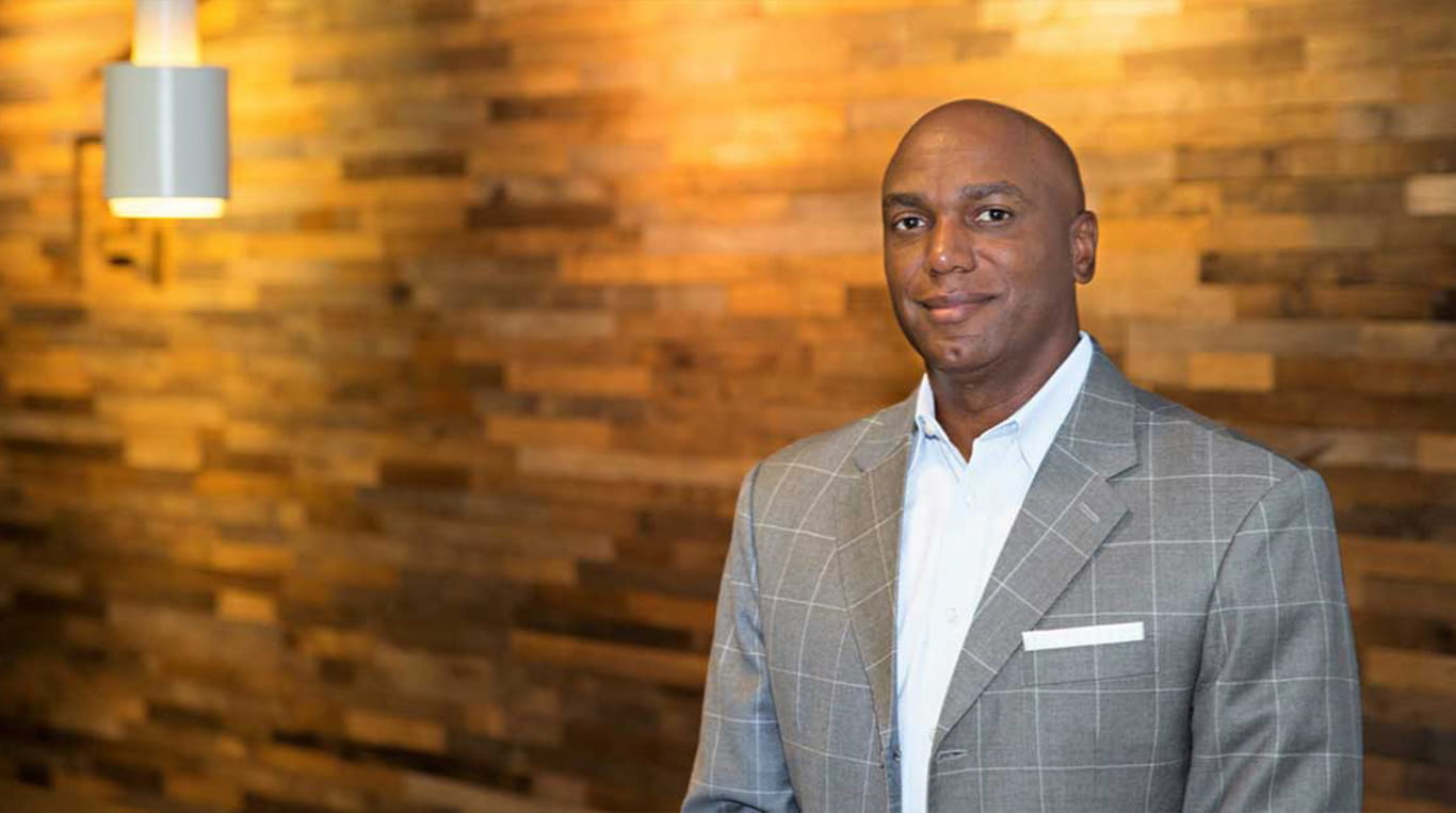 David Nichols, a member of the MBAi Program Industry Advisory Board, wears a gray checkered suit and white shirt, standing in front of a warmly lit, wooden wall.