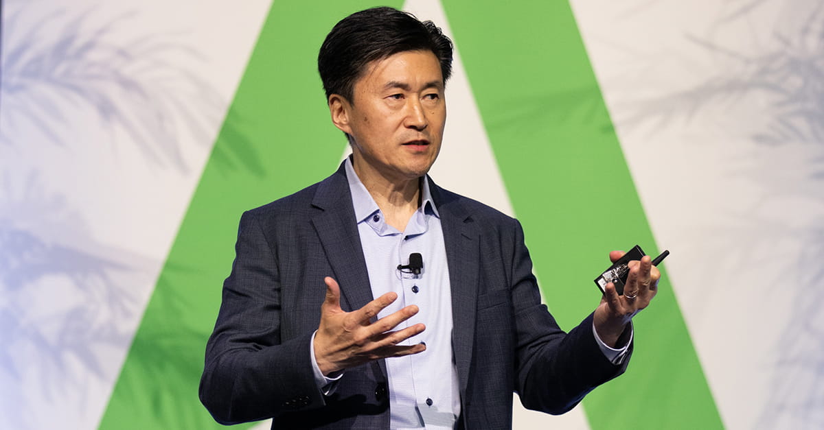 Kellogg alumnus Hoyoung Pak gestures while presenting on stage, holding a remote control device, with a green and white backdrop behind him. He wears a dark blazer and light blue shirt.